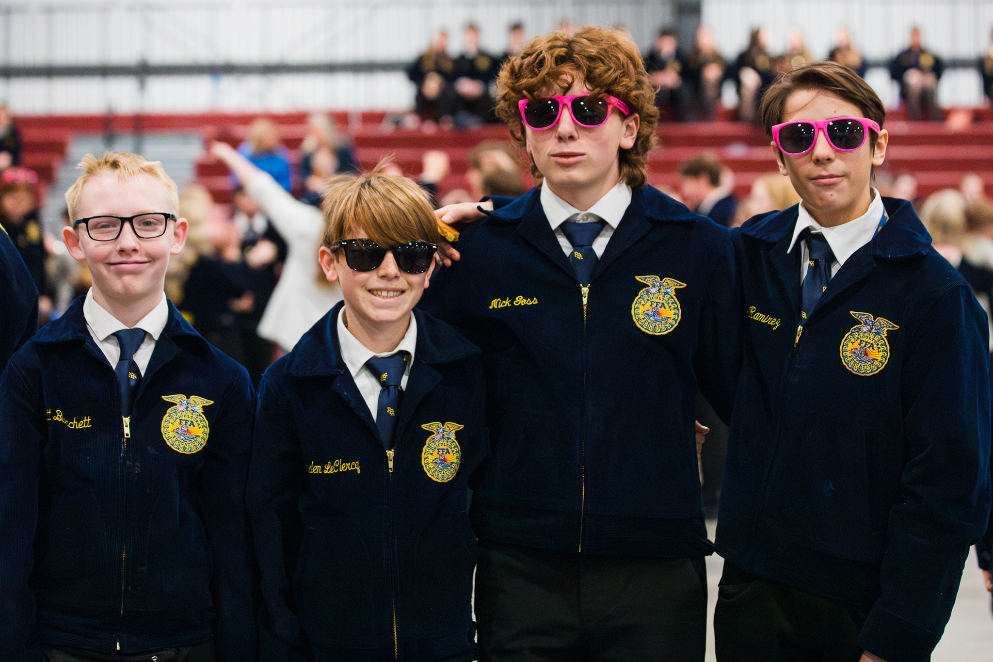 Four FFA members stand together at the state convention wearing official FFA jackets and sunglasses, smiling with other attendees visible in the background.