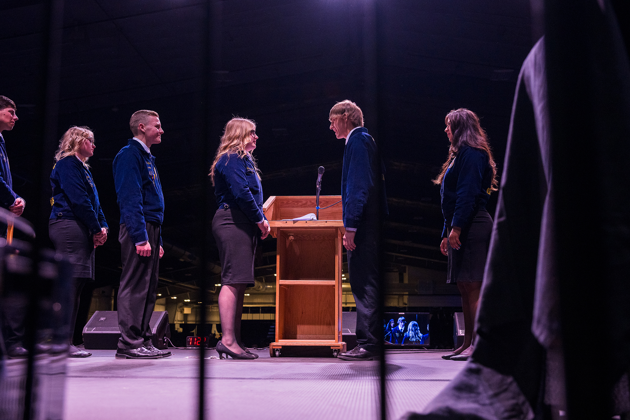 Wyoming FFA state officer candidates stand facing each other on a convention stage behind a podium, dressed in official FFA jackets under dramatic stage lighting during a ceremonial moment.