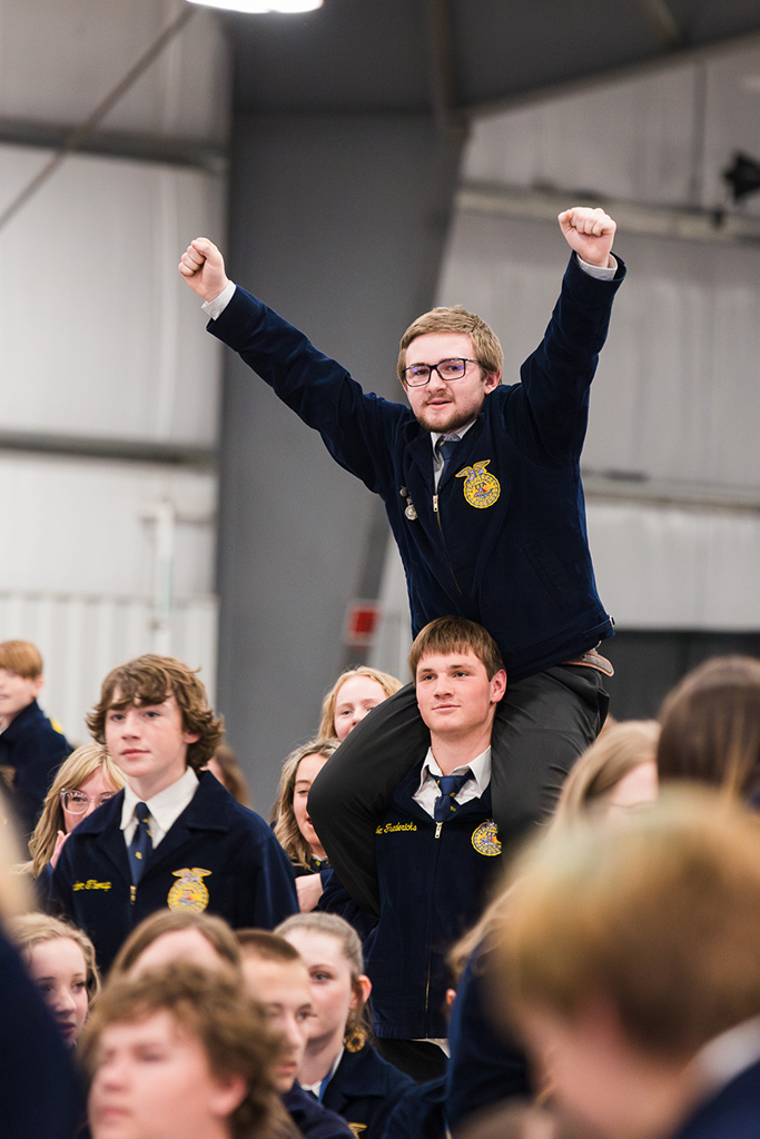 A boy in an FFA jacket with his arms raised sits on the shoulders of another high school age boy.