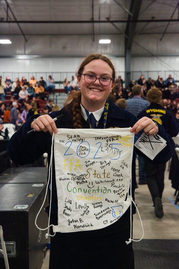 An FFA member smiles while holding a signed white drawstring bag covered with chapter names and messages at the Wyoming FFA State Convention, with other members gathered in the background.