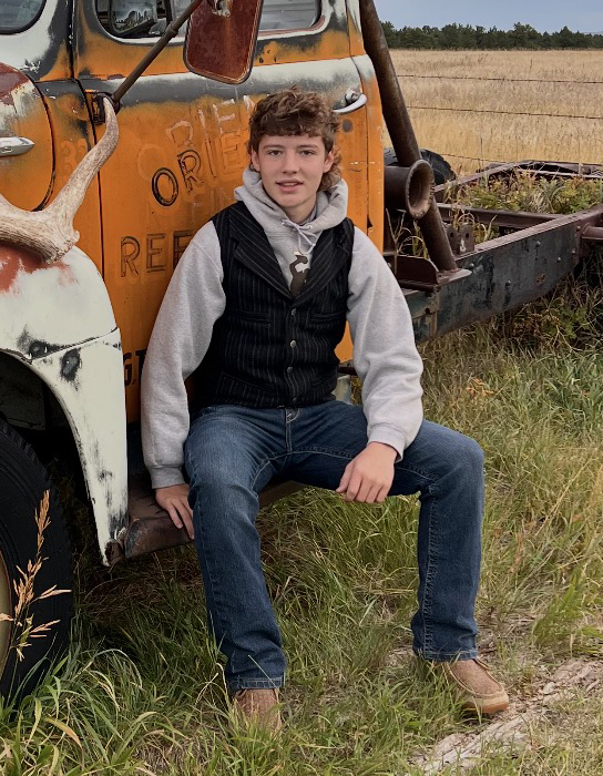 High school student sits on the bumper of a weathered orange farm truck in a rural field.