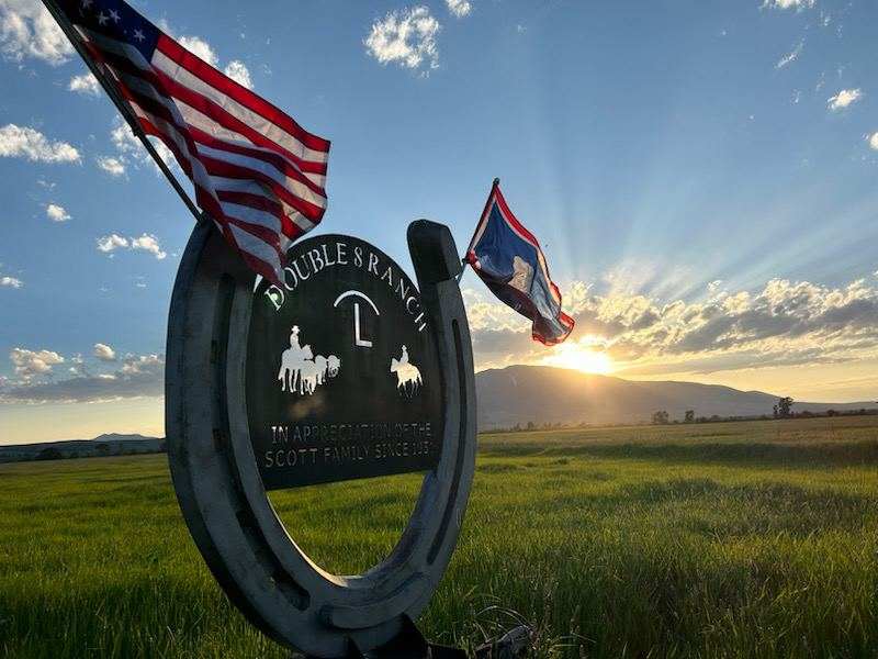 Ranch entrance sign shaped like a horseshoe with American and Wyoming flags flying at sunset over a green pasture.