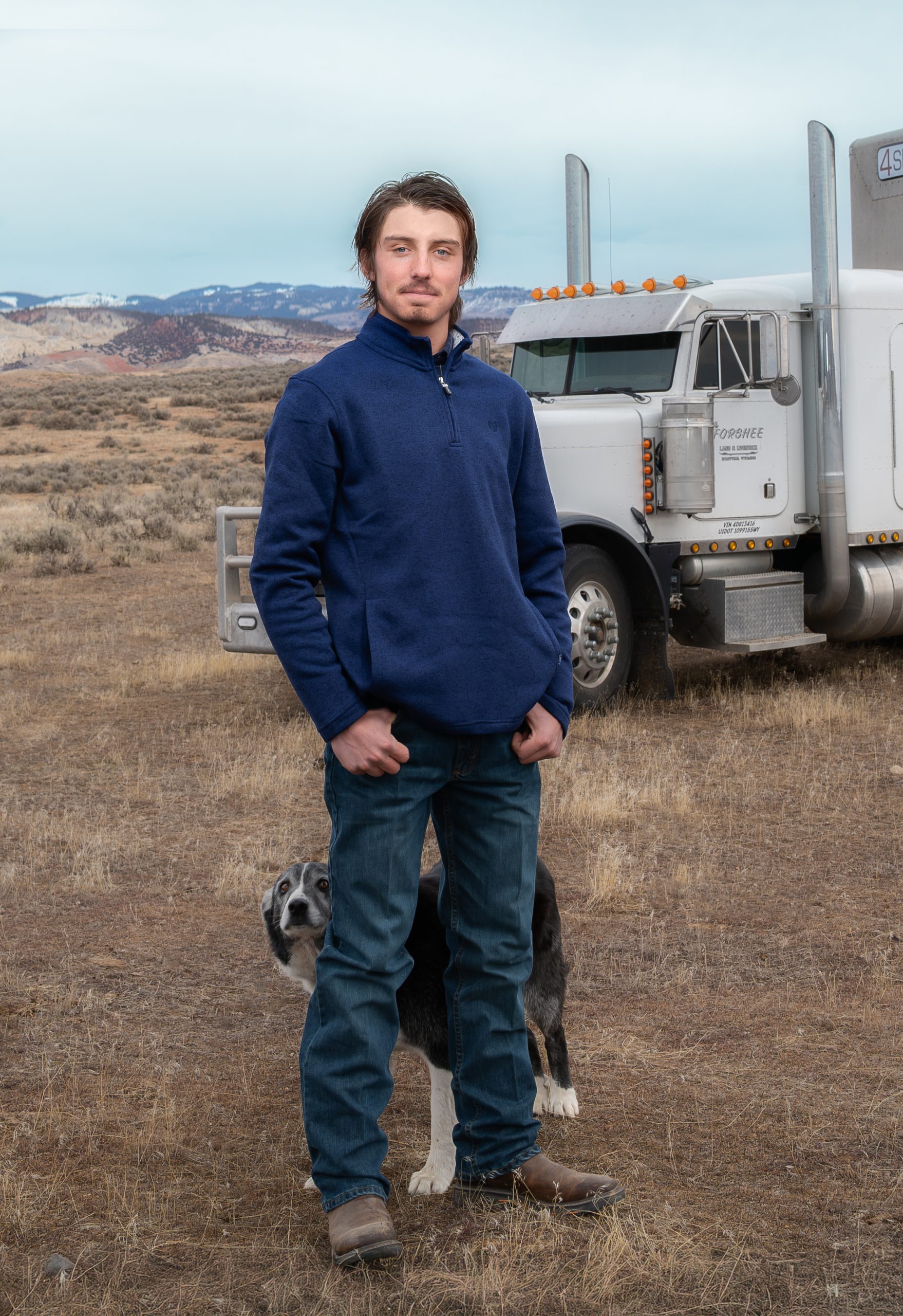 Young rancher standing in Wyoming sagebrush country in front of a semi-truck while his ranch dog sits beside him.